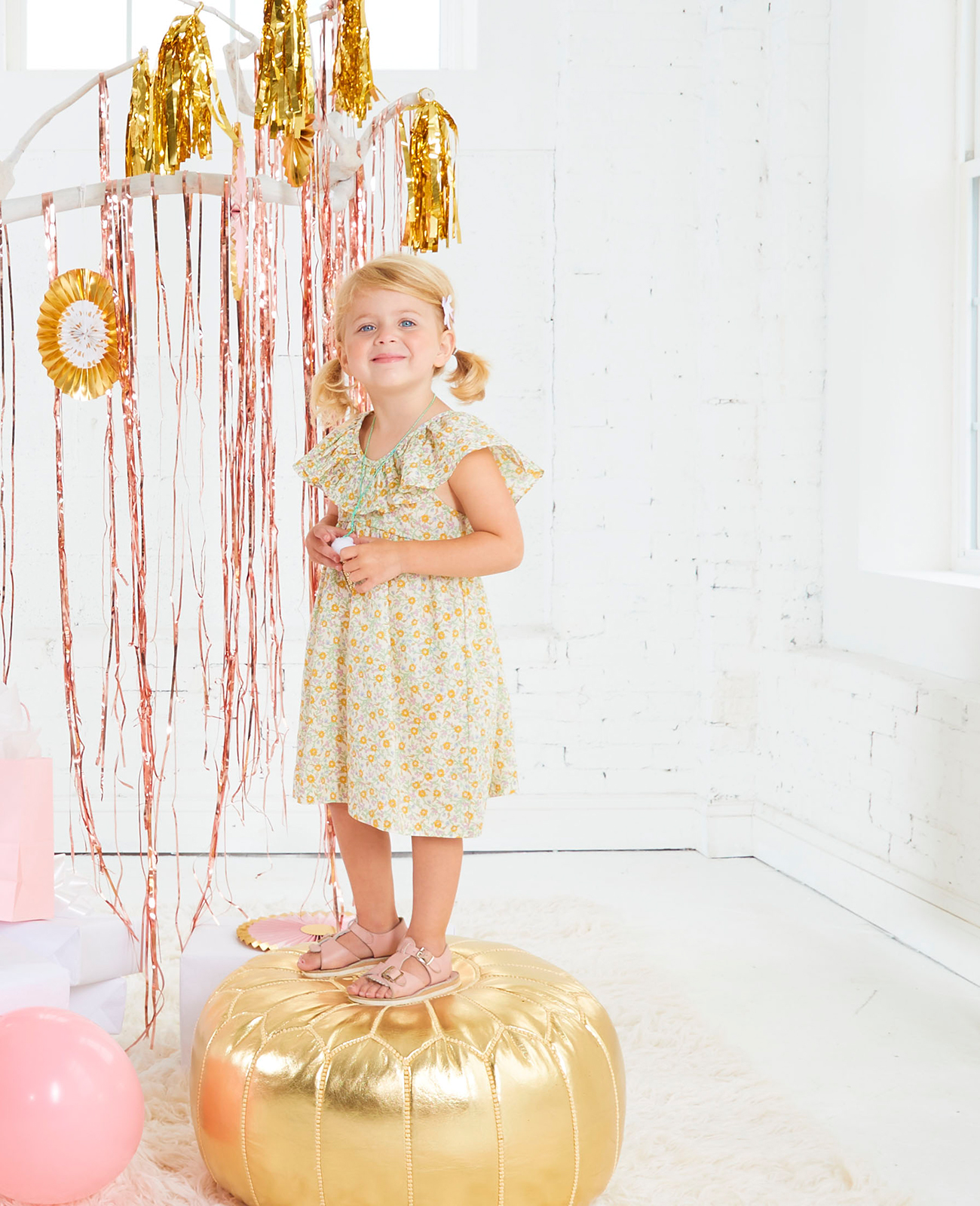 Young girl in a dress standing on gold pouf with gold streamers hanging from white painted tree